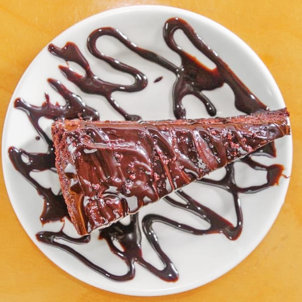 A slice of rich chocolate cake with chocolate drizzle on a white plate.